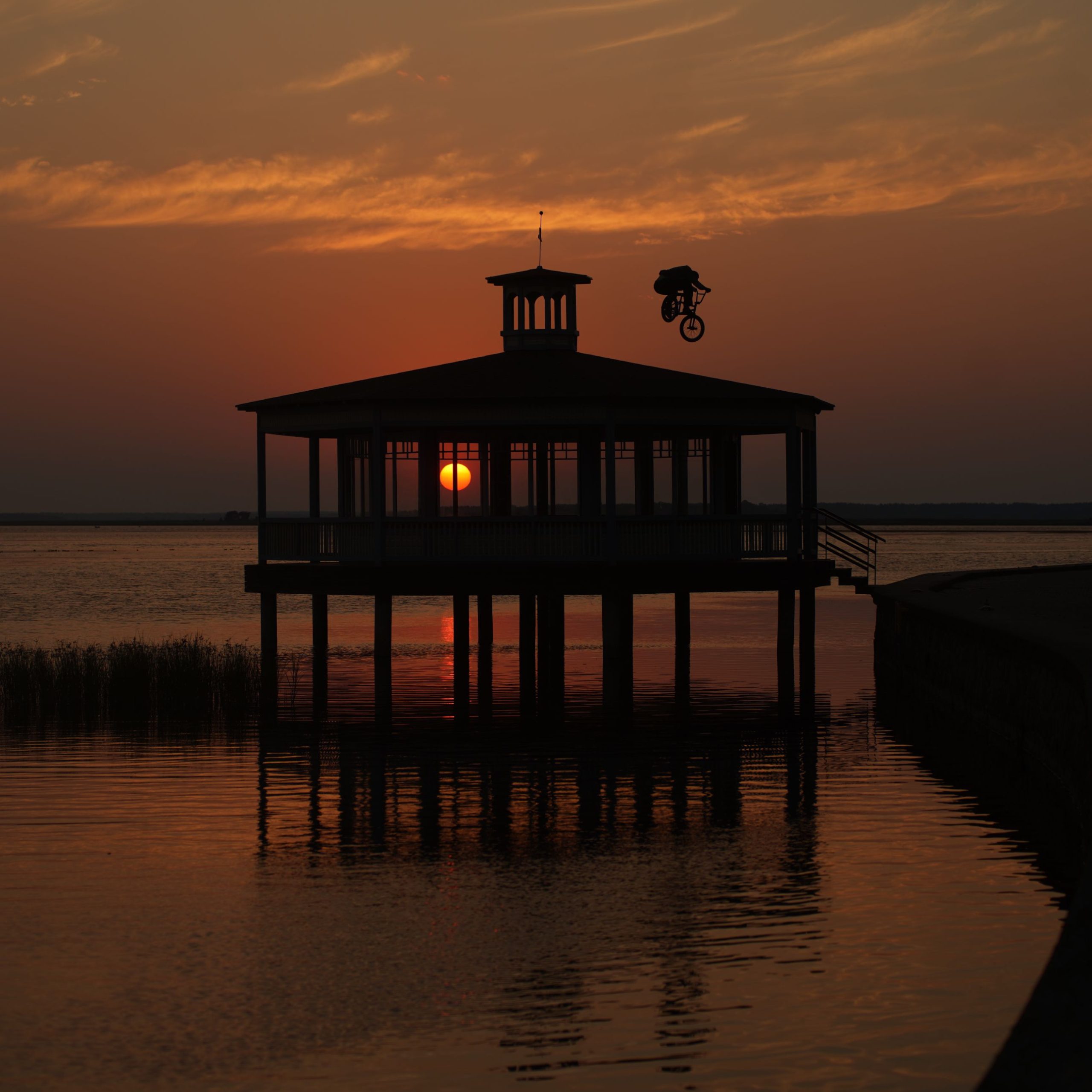 Silhouette of a BMX rider jumping beside a lakeside pavilion at sunset
